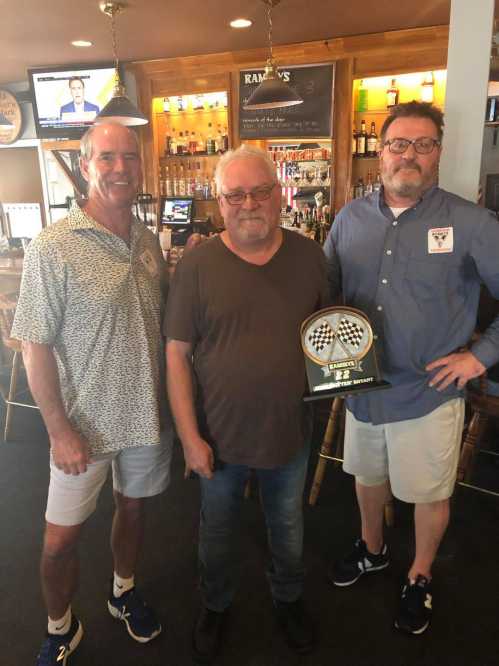 Three men stand together in a bar, one holding a trophy with a checkered flag design.