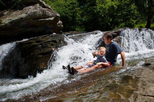 A man and a child sit on a rock by a waterfall, enjoying the splashing water on a sunny day.