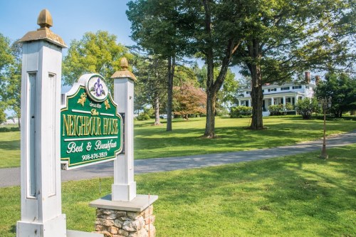 Sign for "The Neighbor House Bed & Breakfast" with a large white house and green lawn in the background.