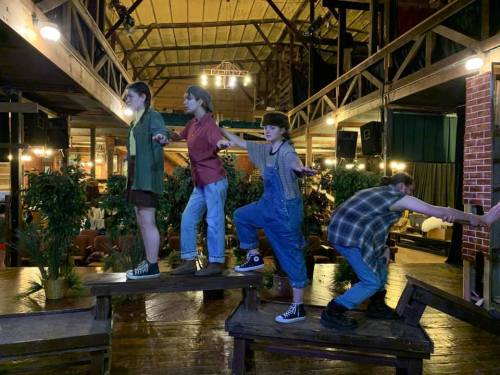 Four performers on a wooden stage, striking dynamic poses amidst greenery and rustic decor in a theater setting.