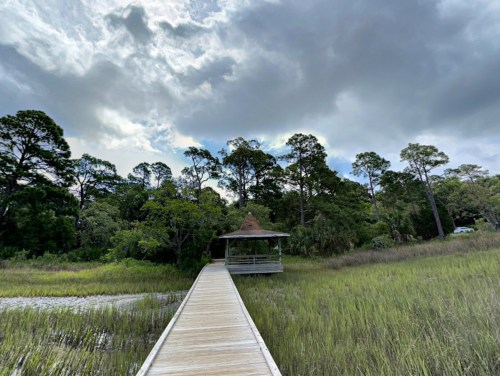 A wooden boardwalk leads to a gazebo surrounded by tall grass and trees under a cloudy sky.