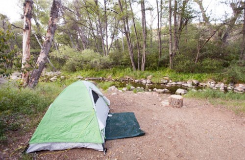 A green tent set up by a peaceful stream, surrounded by trees and rocky terrain.