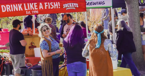 A lively outdoor market scene with people enjoying drinks, colorful tents, and a woman in a fringe vest smiling at the camera.