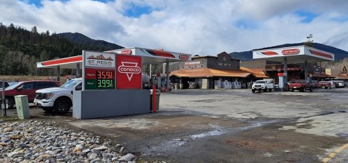 A Conoco gas station with prices displayed, surrounded by vehicles and a rustic store in a mountainous area.