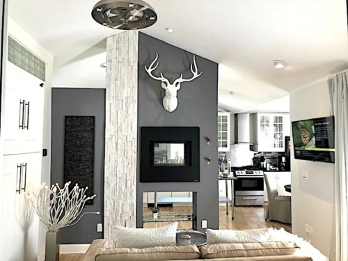 Modern living room with a stone fireplace, antler decor, and a view of a kitchen in the background. Bright and stylish.