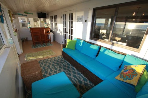 Bright sunroom with turquoise seating, a wicker chair, and a wooden bar, featuring colorful decor and large windows.