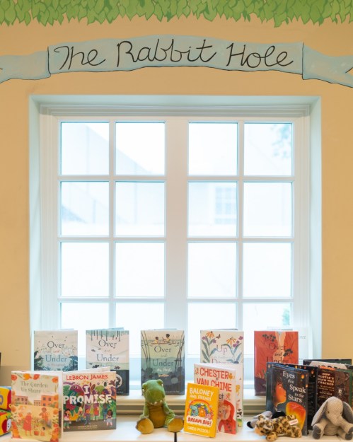 A cozy window display with children's books and plush toys, under a painted sign reading "The Rabbit Hole."