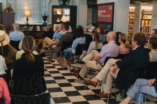 A group of people seated in a library, listening to a speaker at an event, with bookshelves in the background.