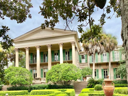 Historic mansion with tall columns, green shutters, and lush gardens, framed by trees under a cloudy sky.