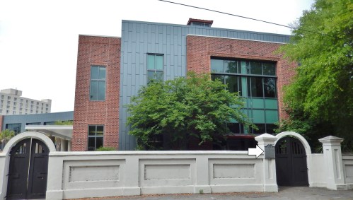 A modern brick building with large windows, surrounded by a white wall and a gate, set in a green landscape.