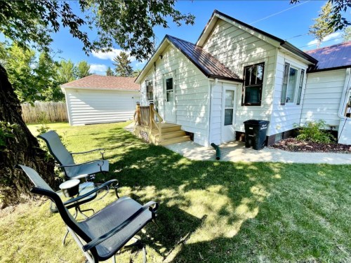 A sunny backyard with two chairs, a small house, and a garage in the background, surrounded by green grass and trees.