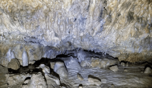 Stalactites and stalagmites in a dimly lit cave, with rocky ground and textured stone walls.