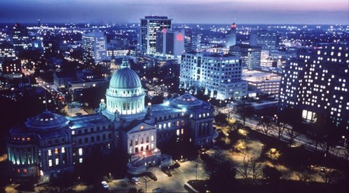 Aerial view of a city skyline at dusk, featuring illuminated buildings and a prominent dome structure.