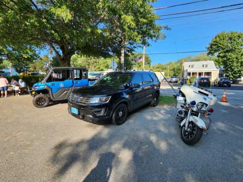 A black SUV, a blue utility vehicle, and a police motorcycle parked on a sunny street near a gathering of people.