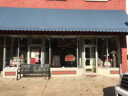 Front view of a storefront with a blue awning, featuring a bench and colorful displays in the windows.