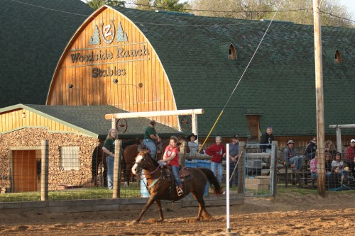 A rider on a horse competes in an arena near Woodside Ranch Stables, with spectators watching from the sidelines.