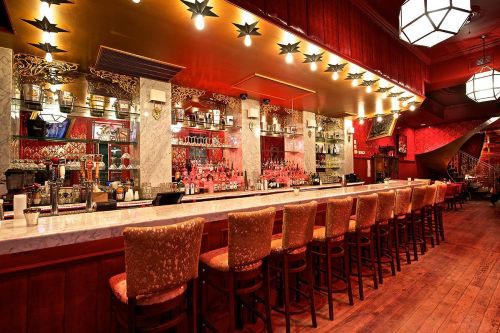 A stylish bar with a long marble counter, high stools, and red decor, featuring shelves of bottles and warm lighting.