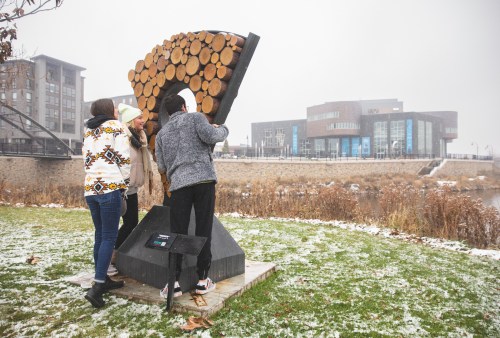 Three people interact with a wooden sculpture in a snowy park, with a modern building visible in the background.