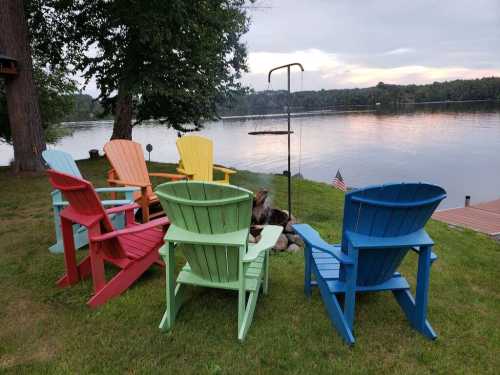 Colorful Adirondack chairs arranged in a circle by a lake, with a fire pit and trees in the background.