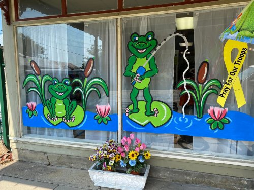 Colorful window mural featuring two frogs fishing on lily pads, surrounded by flowers and a "Pray for Our Troops" ribbon.