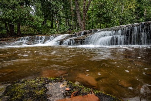 A serene waterfall cascades over rocks into a calm, brown pool, surrounded by lush green trees and foliage.