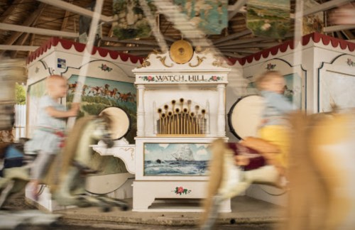 A vintage carousel with a decorative organ and children riding horses, captured in motion.