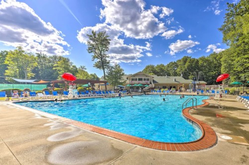 A sunny outdoor pool area with lounge chairs, umbrellas, and people swimming, surrounded by trees and a clubhouse.