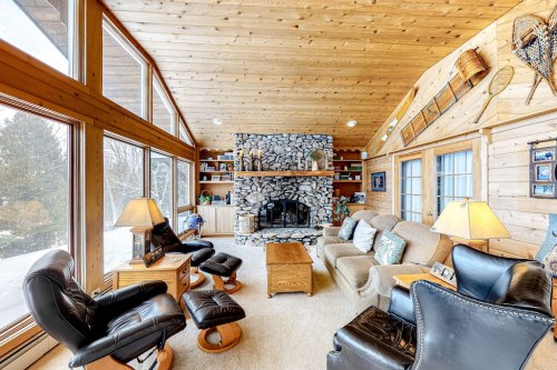 Cozy living room with wooden walls, stone fireplace, large windows, and comfortable seating.