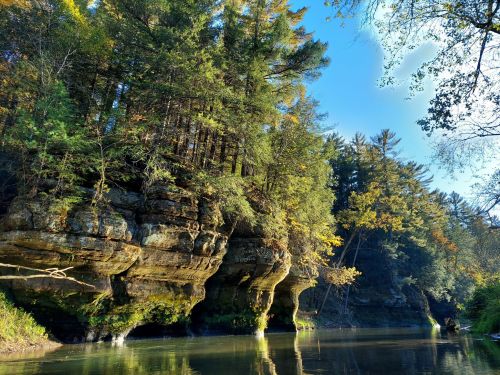 A serene river scene with rocky cliffs and lush trees reflecting in the water under a clear blue sky.