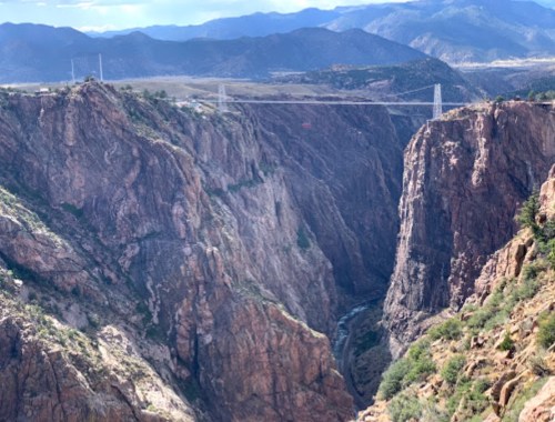 A deep canyon with steep rocky walls and a suspension bridge spanning across it, surrounded by mountains.