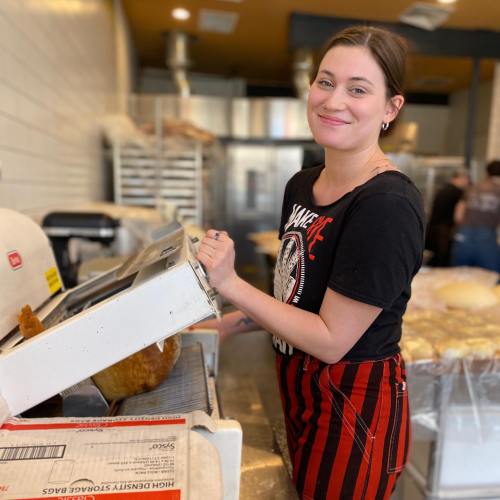 A smiling woman in a black shirt and striped apron operates a bread slicer in a bakery.