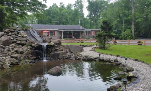 A serene pond with a waterfall, surrounded by rocks and greenery, with a house and patio in the background.