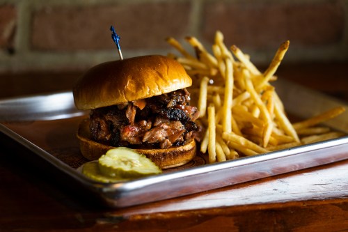 A juicy burger with pulled meat on a bun, served with crispy fries and pickles on a metal tray.