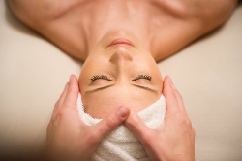 A woman receiving a facial massage, eyes closed, with a towel wrapped around her head. Hands gently apply pressure.