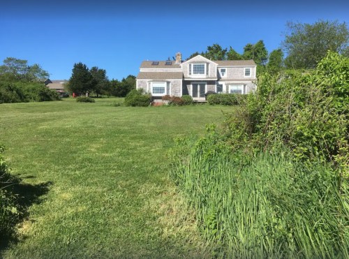 A spacious lawn with a gray house in the background, surrounded by greenery and under a clear blue sky.