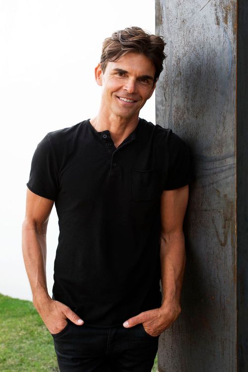 A smiling man in a black shirt stands casually against a metal structure, with greenery and water in the background.