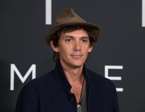 A man with dark hair and a hat poses for a photo against a dark background with text.