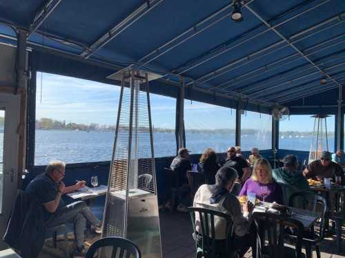 A lively restaurant patio with diners enjoying views of a lake under a blue canopy on a sunny day.