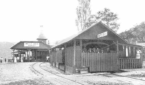 Historic black-and-white photo of a train depot with a wooden structure and people waiting on the platform.