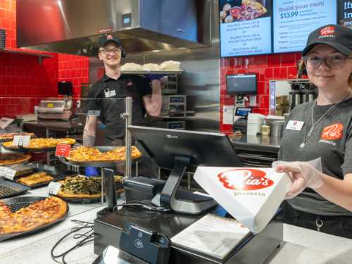 Two employees at a pizzeria smile behind the counter, showcasing pizzas and preparing an order for a customer.