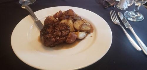 A plate with a cooked steak and roasted potatoes, served with a knife and fork on a dark tablecloth.