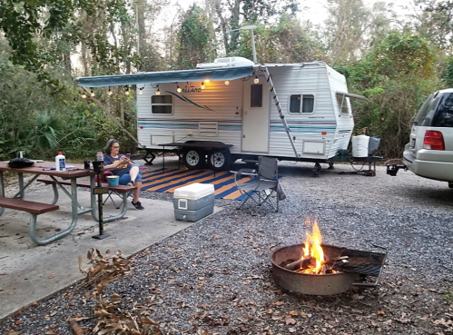 A camper trailer with an awning, picnic table, and a fire pit, surrounded by trees in a serene outdoor setting.