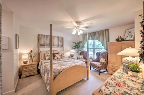Cozy bedroom with a floral bedspread, two armchairs, a ceiling fan, and natural light from a window.