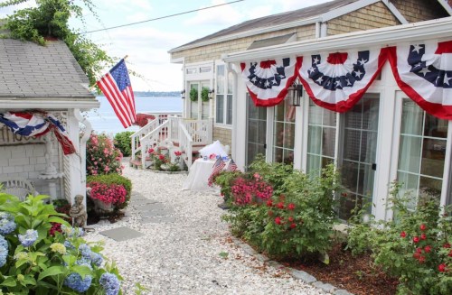 A charming waterfront cottage decorated with red, white, and blue bunting, surrounded by flowers and a stone pathway.