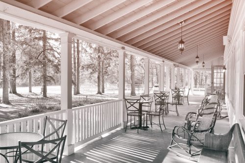 A serene porch with rocking chairs and tables, surrounded by trees and soft sunlight filtering through.
