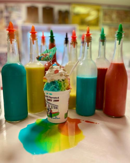 A colorful snow cone topped with whipped cream and a cherry, surrounded by vibrant syrup bottles and a spilled rainbow.