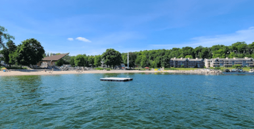 A serene lake view with a floating dock, surrounded by trees and a beach area with people enjoying the sun.