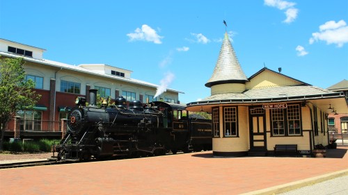 A vintage steam locomotive at a charming train station with a conical roof, set against a clear blue sky.