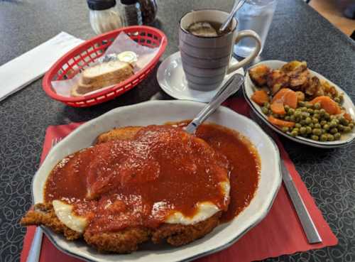 A plate of breaded chicken topped with marinara sauce, served with vegetables, bread, coffee, and a glass of water.