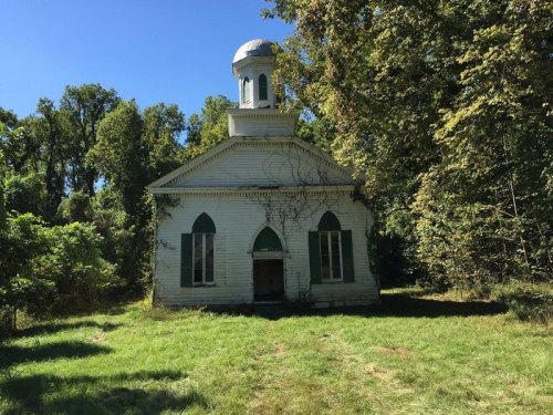 An abandoned white church surrounded by trees and overgrown grass under a clear blue sky.